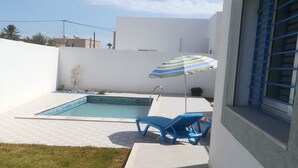 A swimming pool with a tiled deck, a lounge chair, a striped umbrella, and a small tree. - Superb villa near the sea (Houmt Souk)
