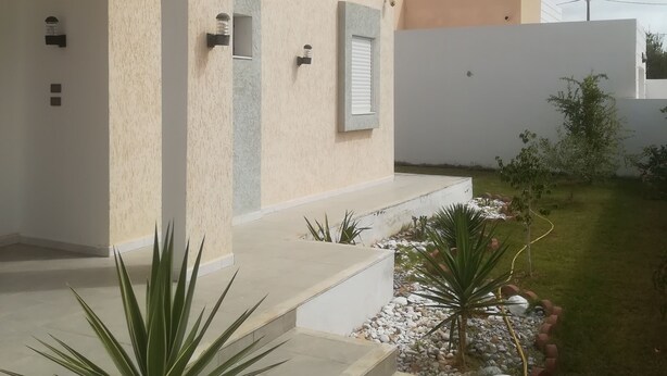 A modern building with a beige exterior, a small garden with plants and a pathway, and a clear sky. - Superb villa near the sea (Houmt Souk)