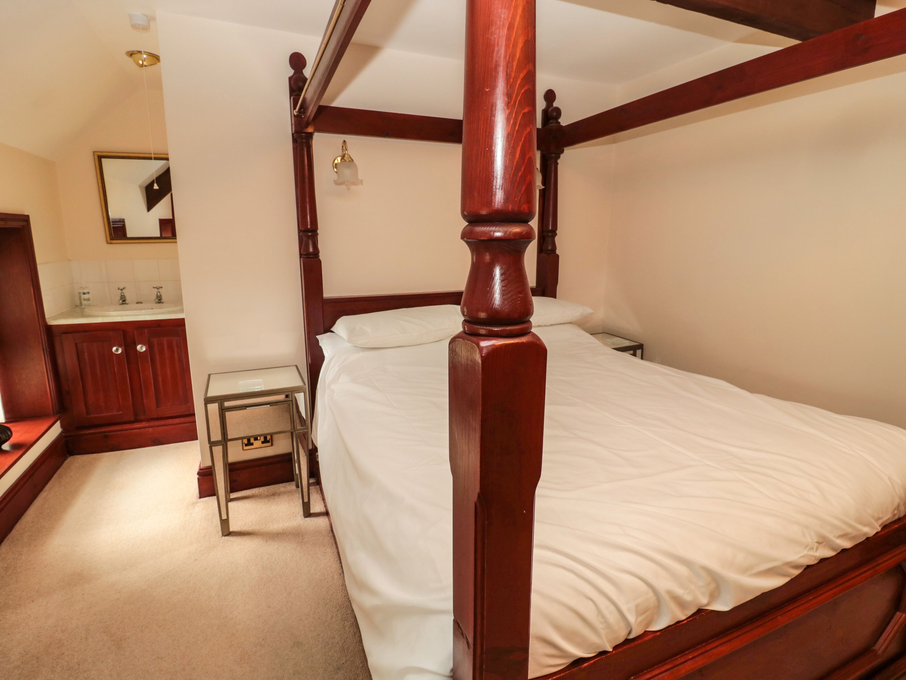 A four-poster bed with white linens, a wooden nightstand, a small table, and a mirror above the sink in a hotel room.