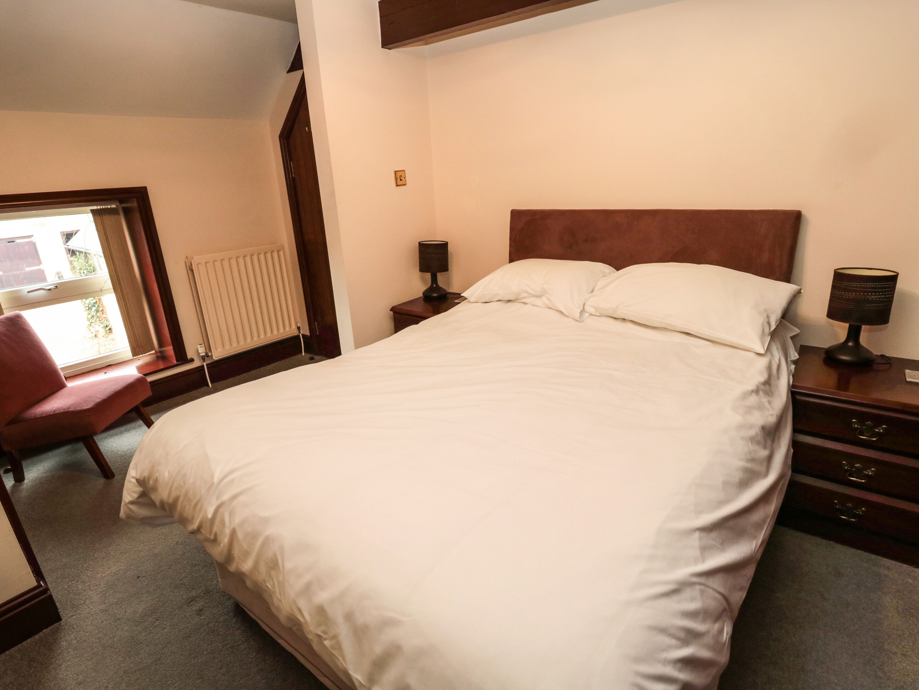 Hotellihuone, jossa on suuri sänky, puinen pääty, yöpöytä lampun kanssa, tuoli ja patteri.