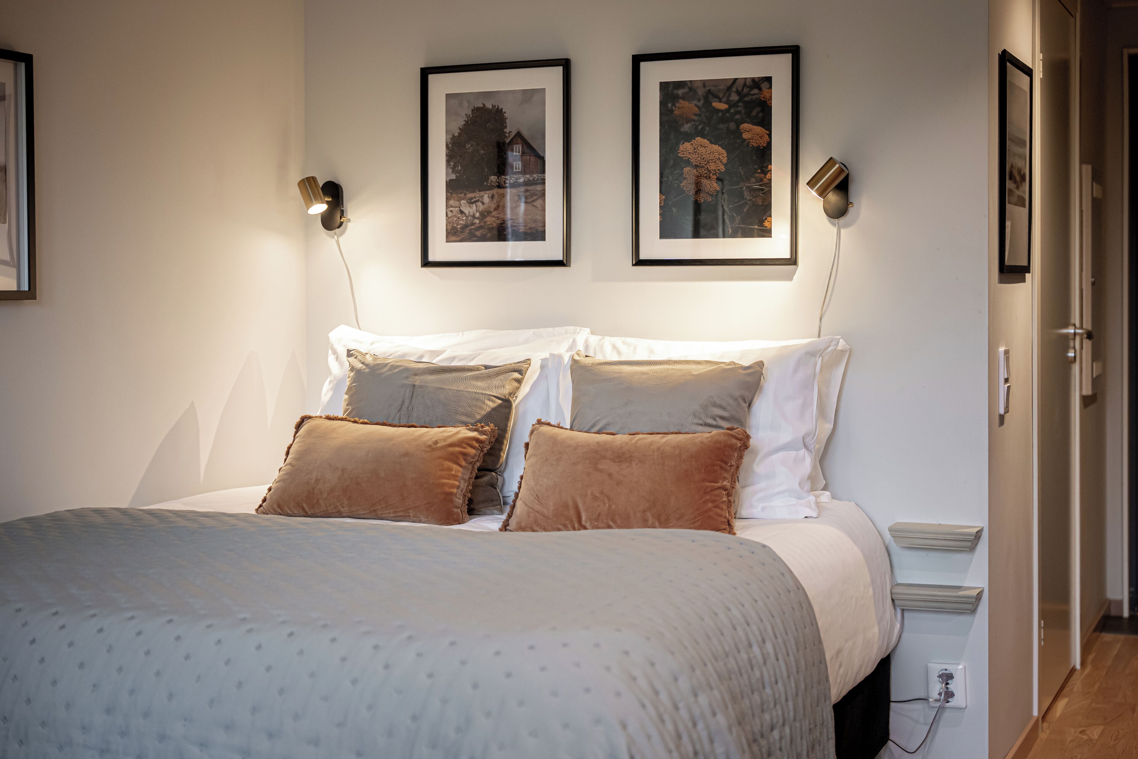 A neatly made bed with a patterned bedspread, two framed pictures on the wall, and a bedside table with a lamp.