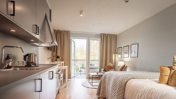 Una camera d'albergo moderna con angolo cottura, un letto con copriletto color beige, un tavolino e vista sul balcone esterno.