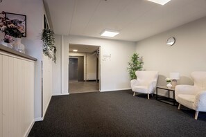 Un couloir doté d’un sol recouvert de moquette, de murs blancs et d’une porte donnant sur une autre pièce. On y trouve deux chaises blanches et une petite table avec une lampe.