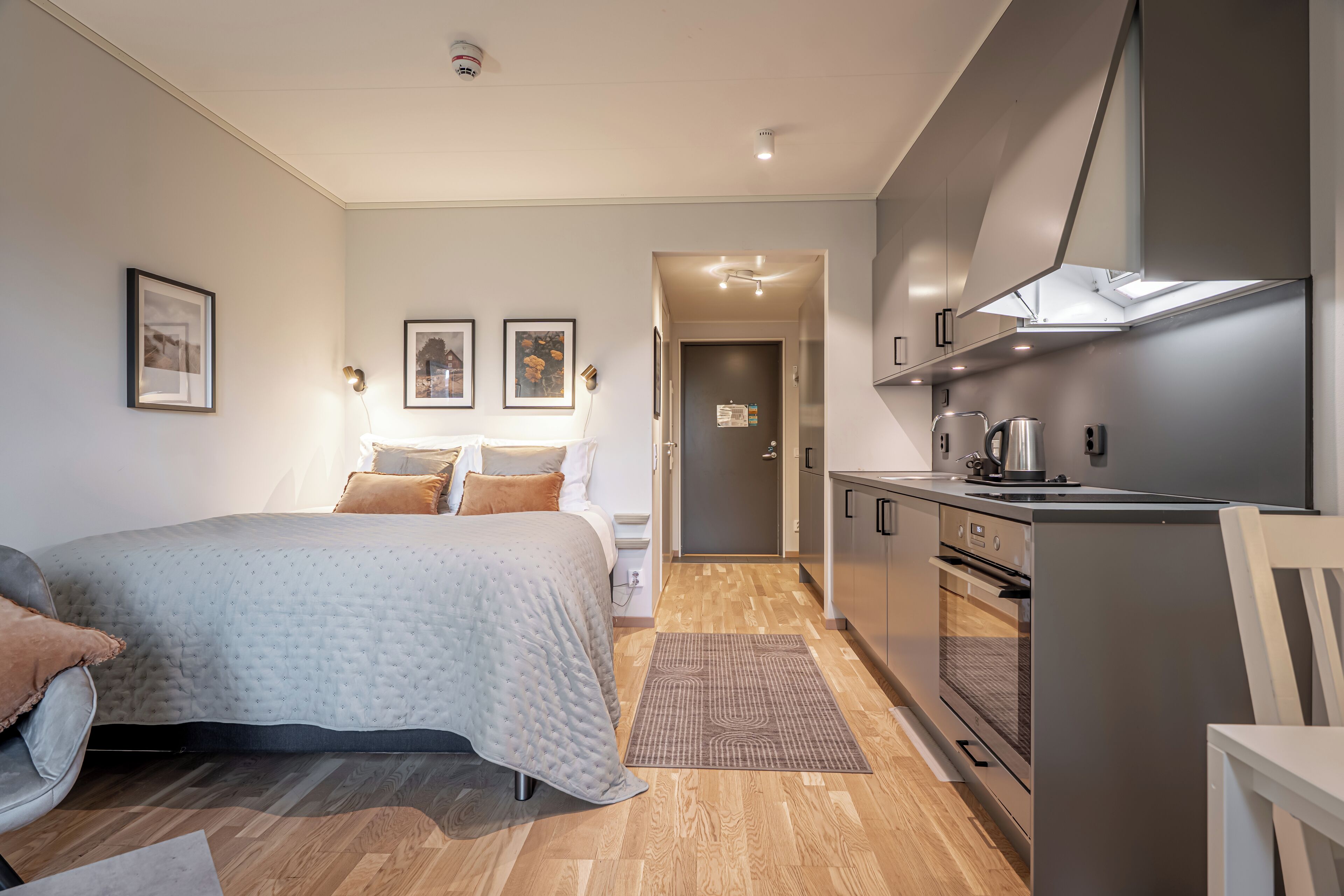 Una habitación de hotel moderna con cama, mesita de noche, lámpara y una zona de kitchenette.