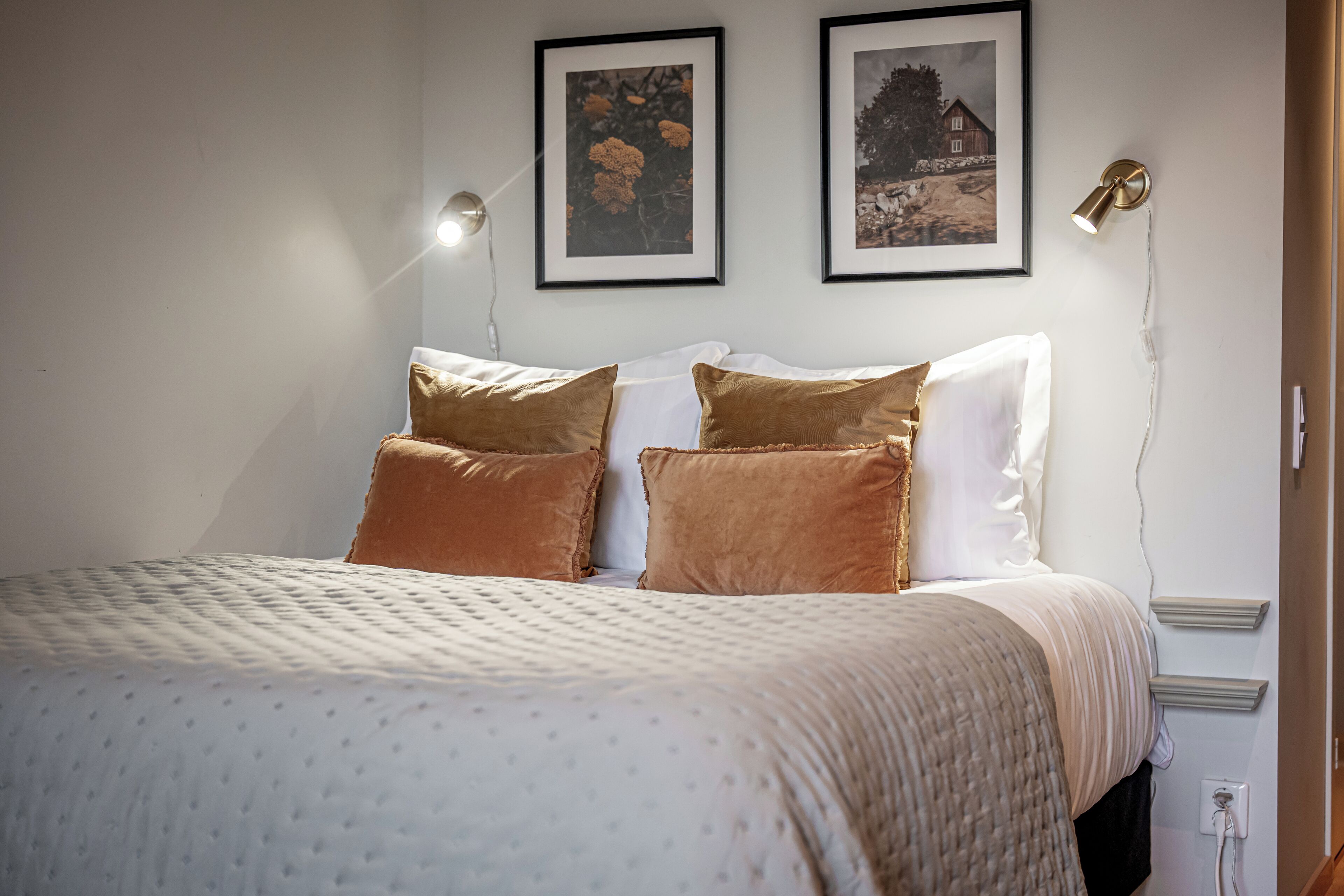 A neatly made bed with a patterned bedspread and two pillows, a wall-mounted lamp, and two framed pictures above the bed.