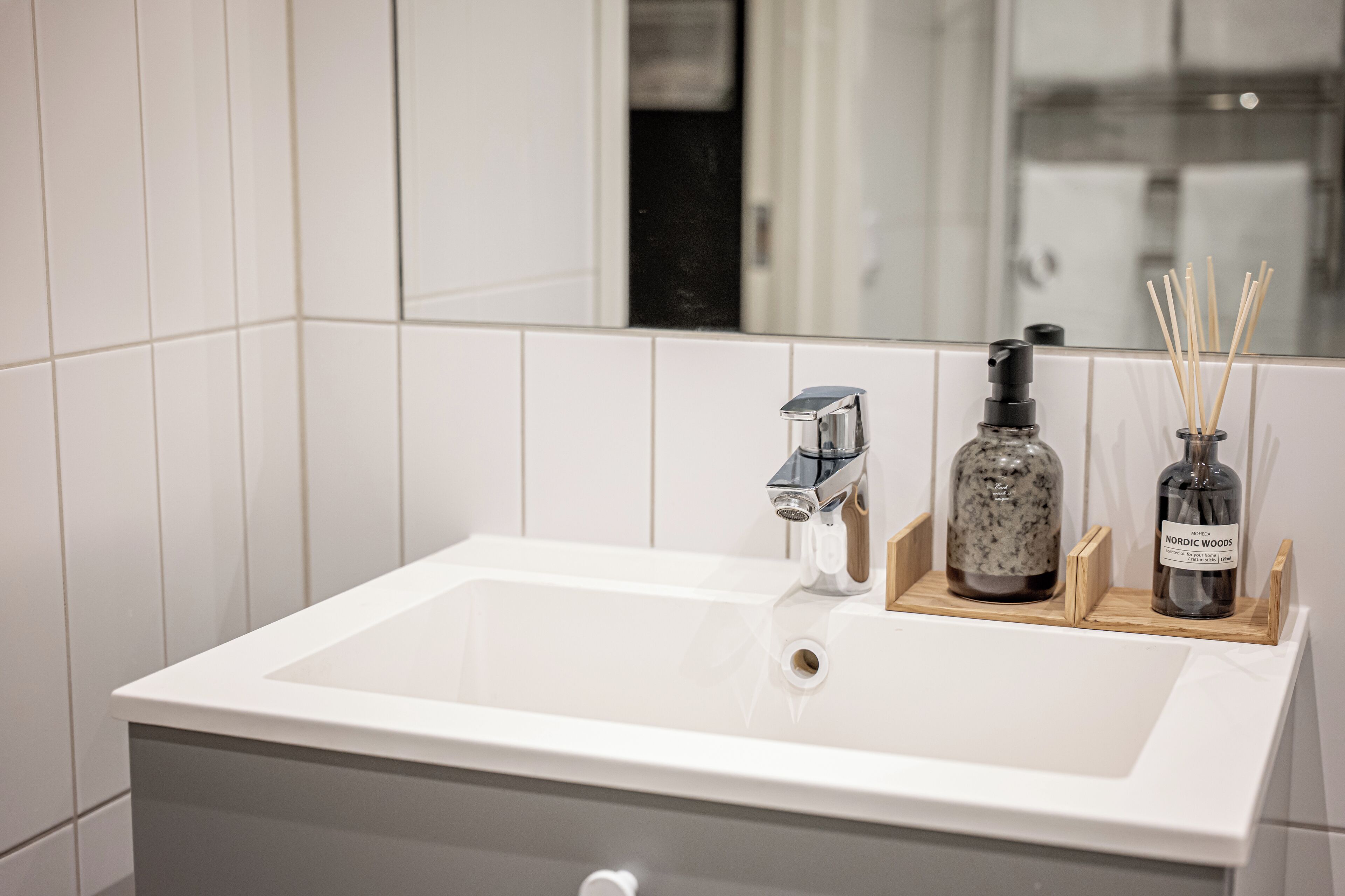 Un baño con un lavamanos blanco, un espejo y una bandeja de madera con una botella y difusor de cañas.