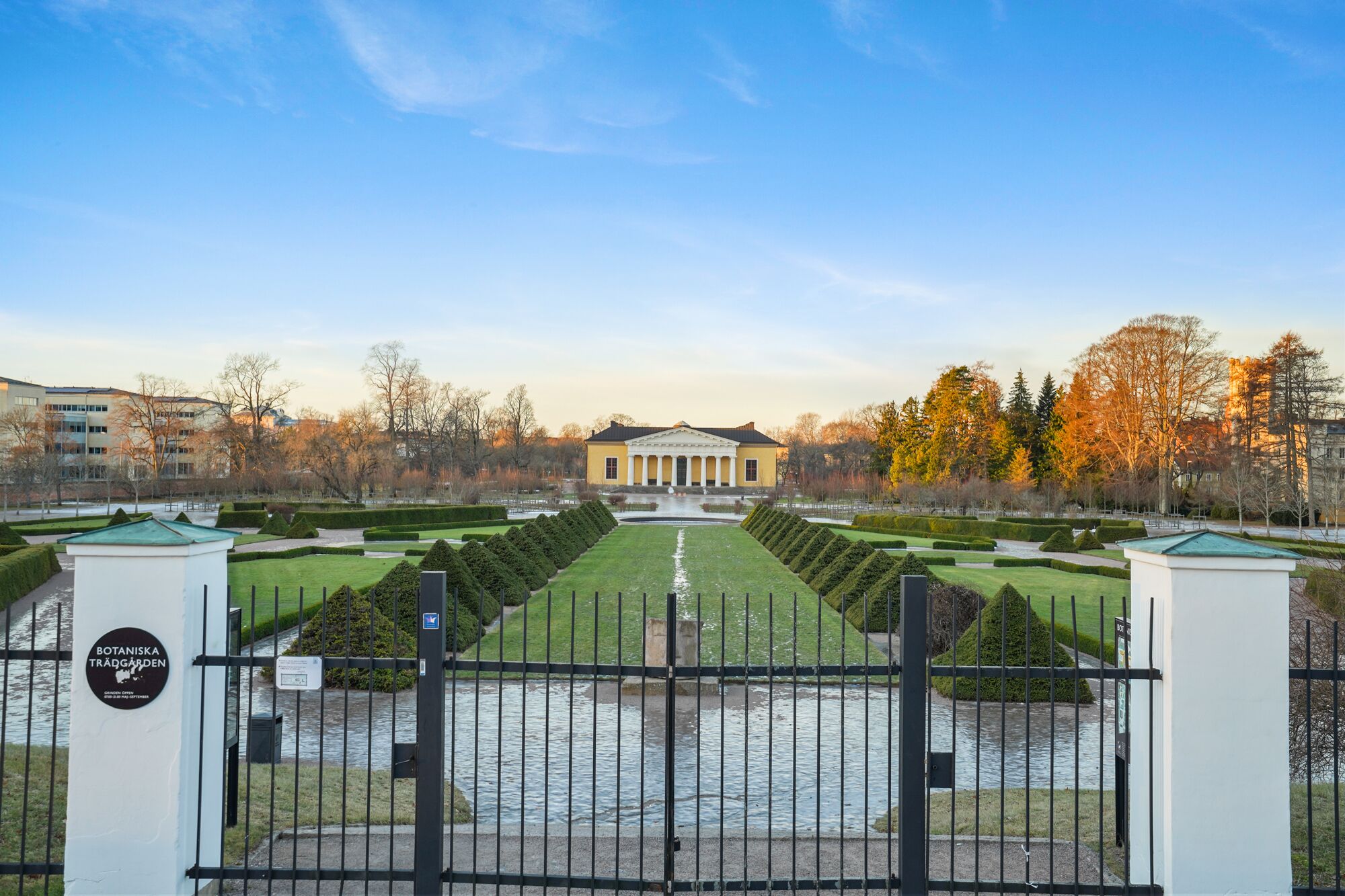 A formal garden with a symmetrical layout, a central water feature, and a classical building in the background.