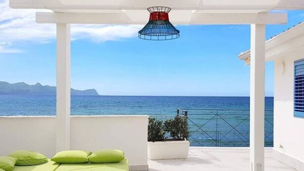 Ein Balkon mit weißer Struktur, grünen Kissen, einem Topfpflanzen und Blick aufs Meer.