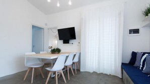 Ein modernes Zimmer mit Esstisch, Stühlen, Fernseher, Bett und einem Fenster mit Vorhängen.