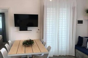 Ein Zimmer mit einem Holzespeisetisch, weißen Stühlen, einem an der Wand befestigten Flachbildfernseher und einem Fenster mit transparenten Vorhängen.