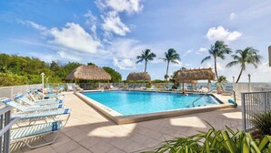 A swimming pool with lounge chairs and thatched-roof structures, surrounded by palm trees and a clear sky.