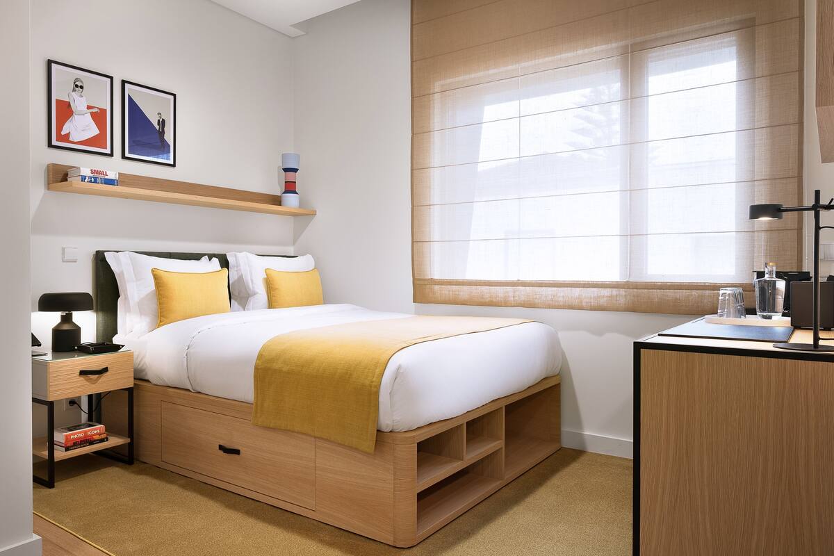 A neatly arranged bedroom with a bed, bedside table, lamp, and a desk with a lamp.