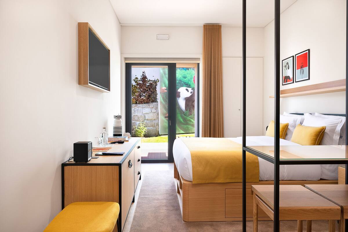 A modern hotel room with a bed, a desk, and a seating area. There is a sliding door leading to a balcony with a view of greenery.