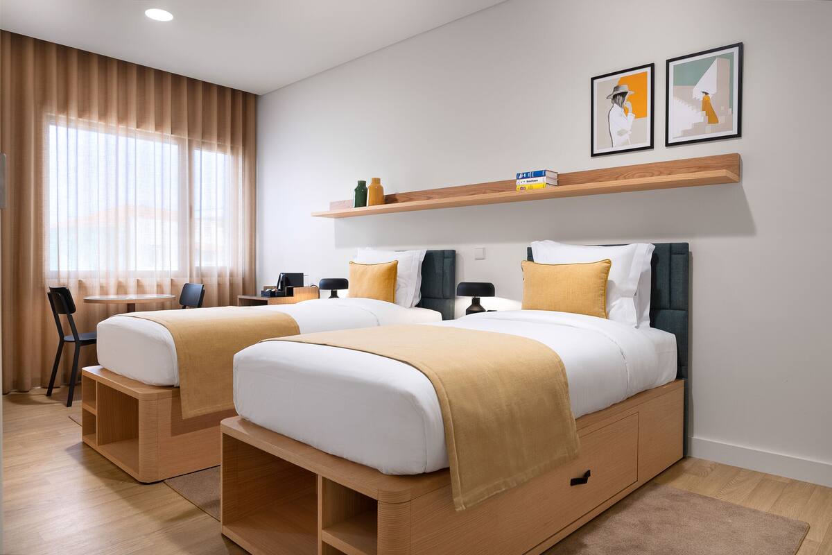 A hotel room with two beds, a desk, a chair, a shelf with books and bottles, and two framed pictures on the wall.