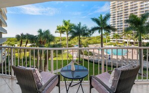 A balcony with wicker furniture and a glass table, overlooking a pool area with palm trees and a hotel building in the background.