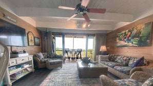 A spacious living room with a large TV, wicker furniture, a ceiling fan, and a view of the outdoors through large windows.
