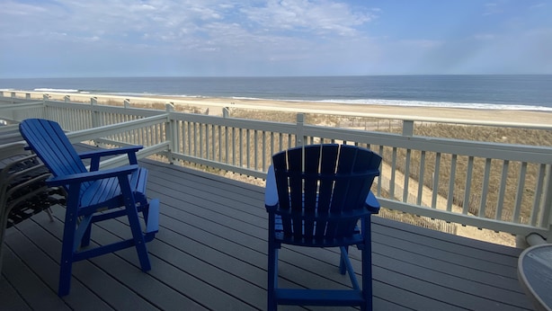 A wooden deck with two blue Adirondack chairs facing a beach.