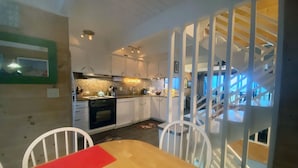 A dining area with a wooden table and chairs, a kitchen in the background, and a staircase with a white railing.