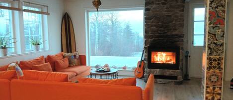 A living room with a stone fireplace, orange sofas, and a view of trees through large windows.