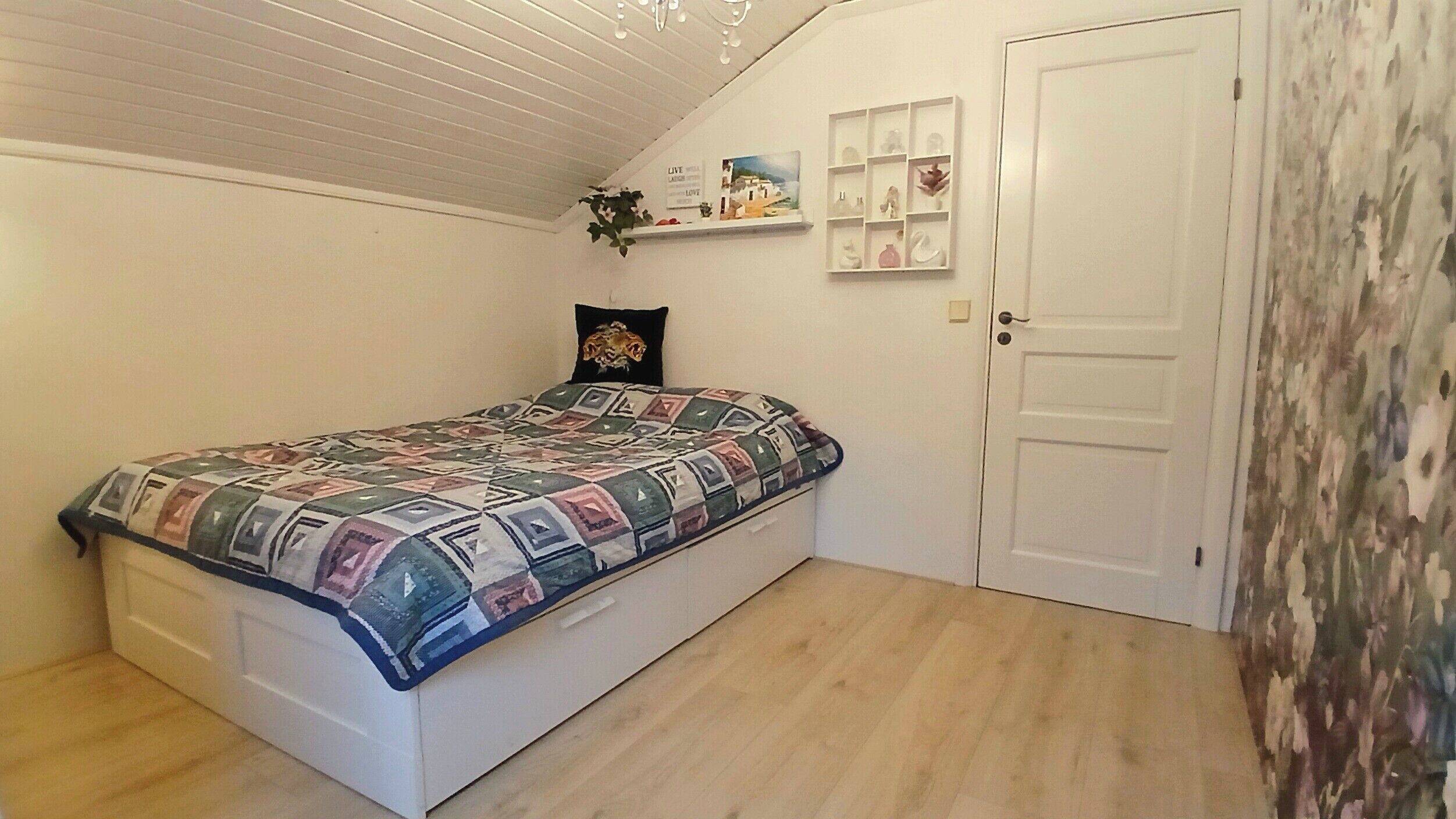 A bedroom with a bed, a door, a shelf with decorative items, and a wall with a floral pattern.