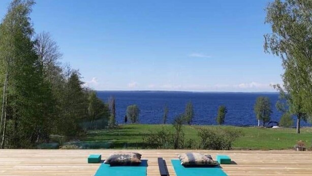 Una terraza de madera con dos esterillas y almohadones azules, con vistas a un cuerpo de agua y vegetación.