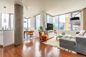 A modern living room with a grey sofa, a dining area with orange chairs, a TV on a white cabinet, and large windows offering city views. - 2BR | Gym, Pool | Bellevue | Sophari Towers L (Bellevue)