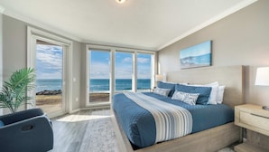Une chambre à coucher avec un grand lit, une table de chevet, une chaise et une vue sur l’océan grâce à de grandes fenêtres.