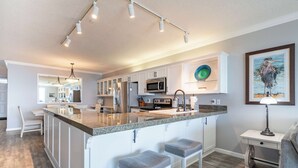 Une cuisine moderne avec des meubles blancs, un comptoir en granit et des appareils en acier inoxydable. On y trouve deux tabourets de bar et une petite table avec une lampe.