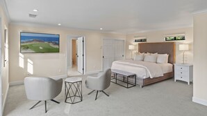 A modern bedroom with a large bed, two chairs, a small table, a TV, and a door leading to another room.