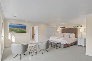 Ein modernes Schlafzimmer mit einem großen Bett, zwei Stühlen, einem kleinen Tisch, einem Fernseher und einer Tür zu einem anderen Raum.