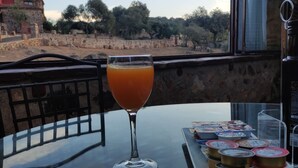 Une table avec un verre de jus d’orange, une tasse de café et plusieurs récipients contenant de la nourriture, le tout placé sur un balcon donnant sur un mur de pierre et des arbres.