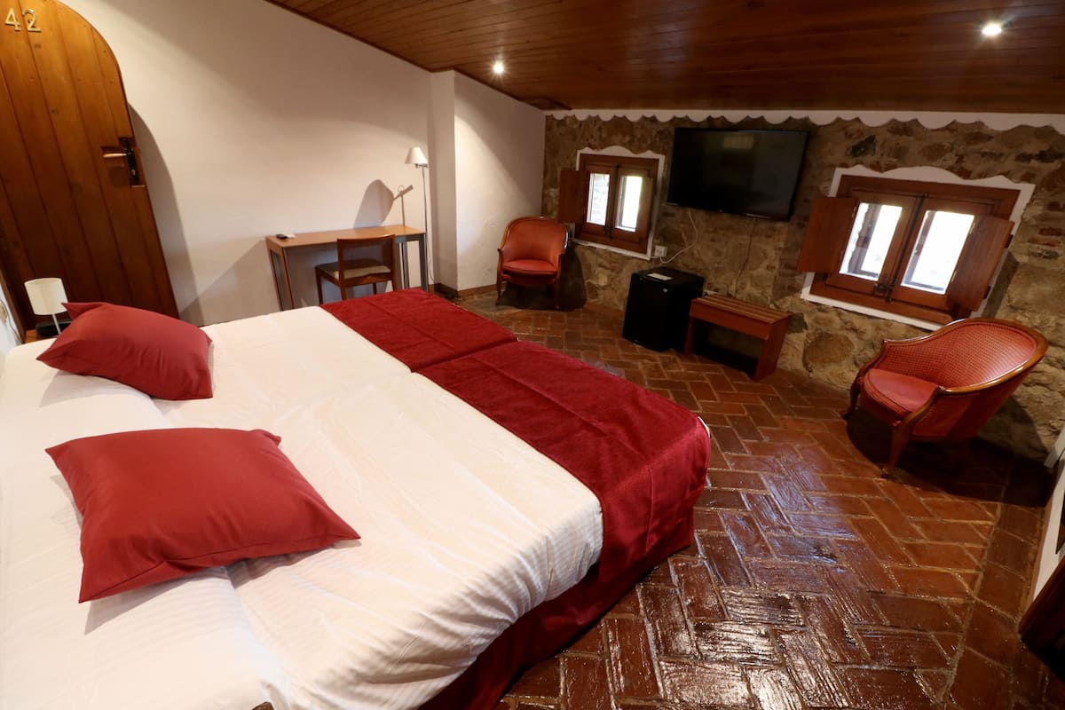 Une chambre à coucher avec un grand lit, deux fauteuils rouges, un bureau en bois et un téléviseur fixé à un mur en pierre.