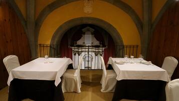 A dining area with two tables set for two, each with a white tablecloth and black chair covers. The room features a vaulted ceiling with a decorative emblem above the tables.