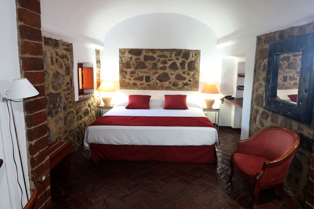 Une chambre avec un mur en pierre, un lit avec une literie rouge, une chaise en bois et une cheminée en pierre.