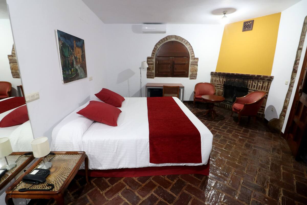 Une chambre d’hôtel avec un lit, des oreillers rouges, une petite table avec une lampe, une chaise et une cheminée.