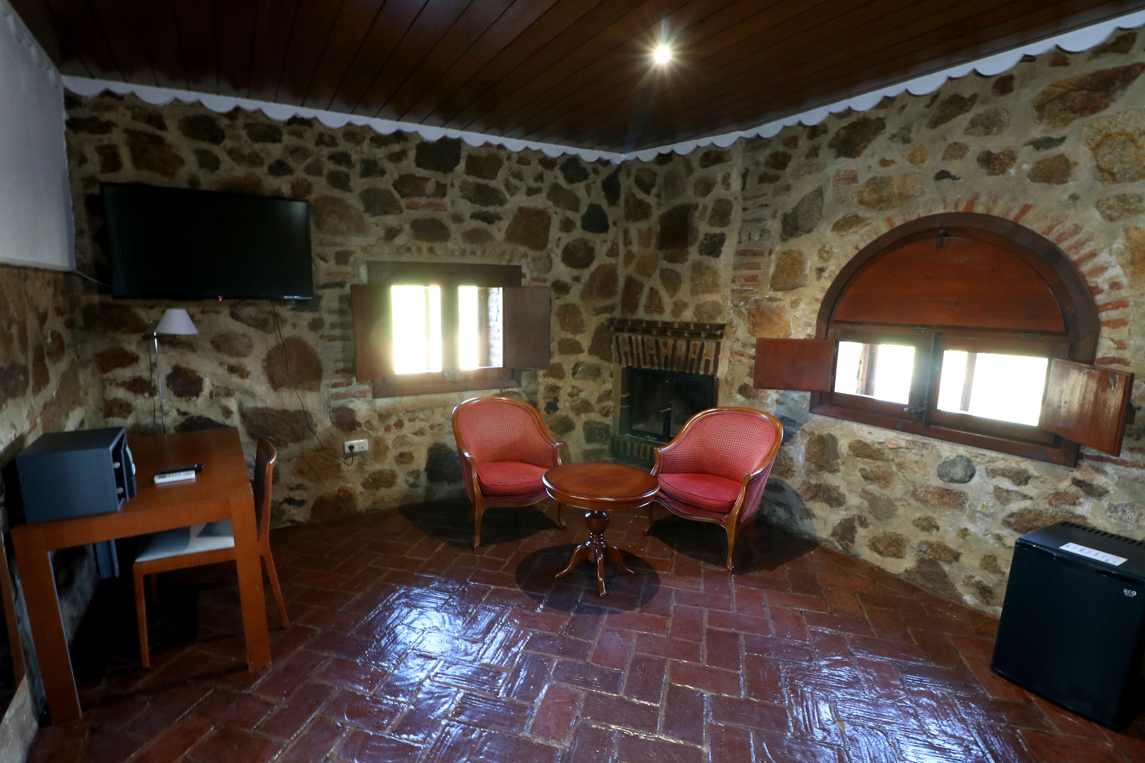 A stone-walled room with a TV, two red chairs, a small table, and a fireplace.
