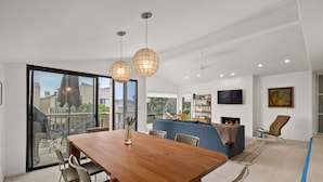 A modern living room with a wooden dining table, two pendant lights, a blue sofa, a TV, and a fireplace. - Hacienda Palms - Vacay for 6 - Near Ocean, Pools, Tennis, Racetrack (Solana Beach)