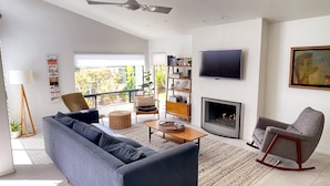 A modern living room with a grey sofa, a wooden coffee table, a fireplace, a television, and a bookshelf. - Hacienda Palms - Vacay for 6 - Near Ocean, Pools, Tennis, Racetrack (Solana Beach)