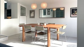 Un espace de salle à manger moderne avec une table en bois et des chaises en métal, des luminaires suspendus, un grand miroir et des œuvres d’art encadrées.