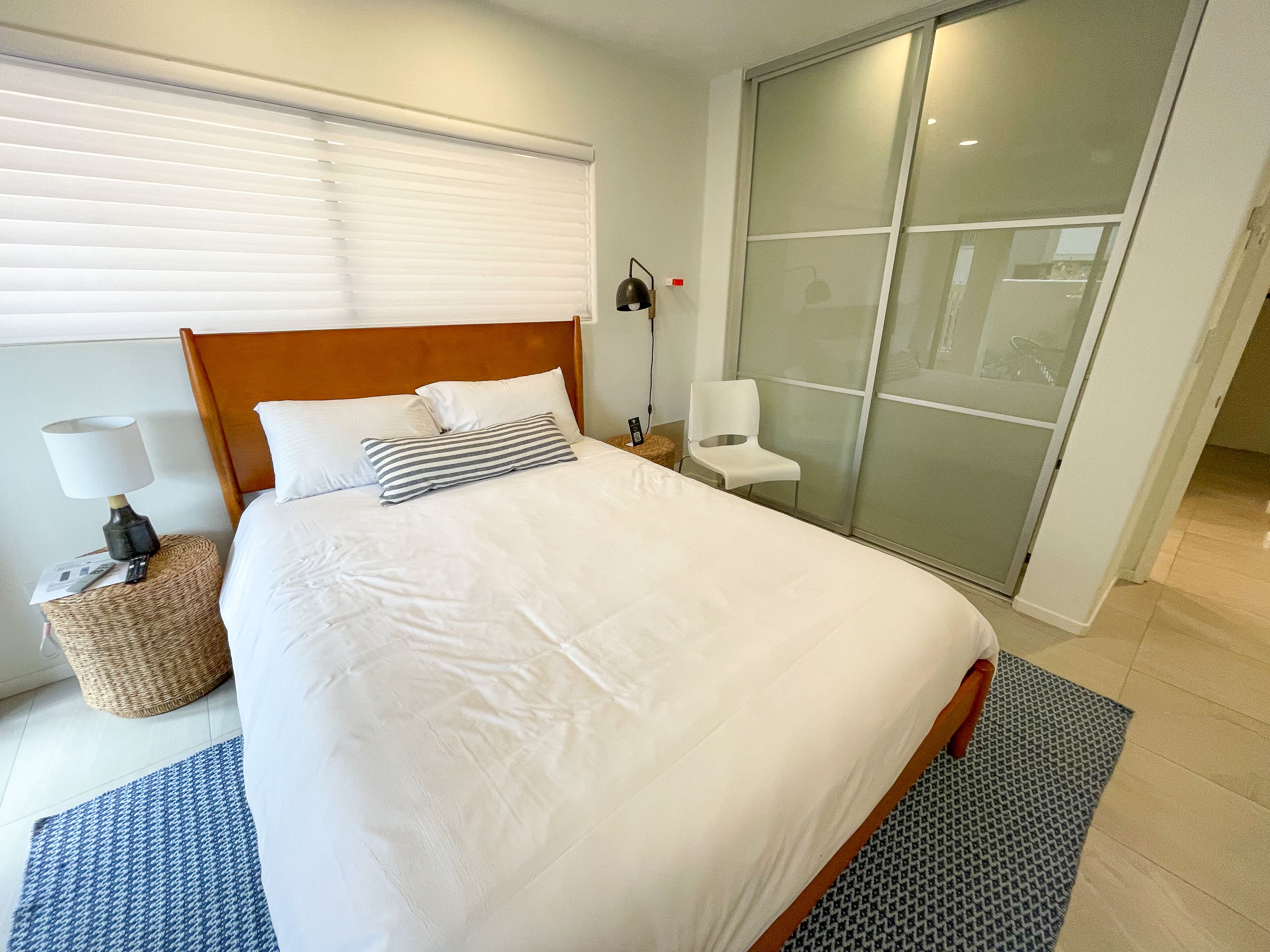 Un lit bien fait, avec une literie blanche, une tête de lit en bois et un coussin à rayures. On aperçoit une lampe sur une table de chevet, une chaise et, au fond, une porte-fenêtre coulissante.