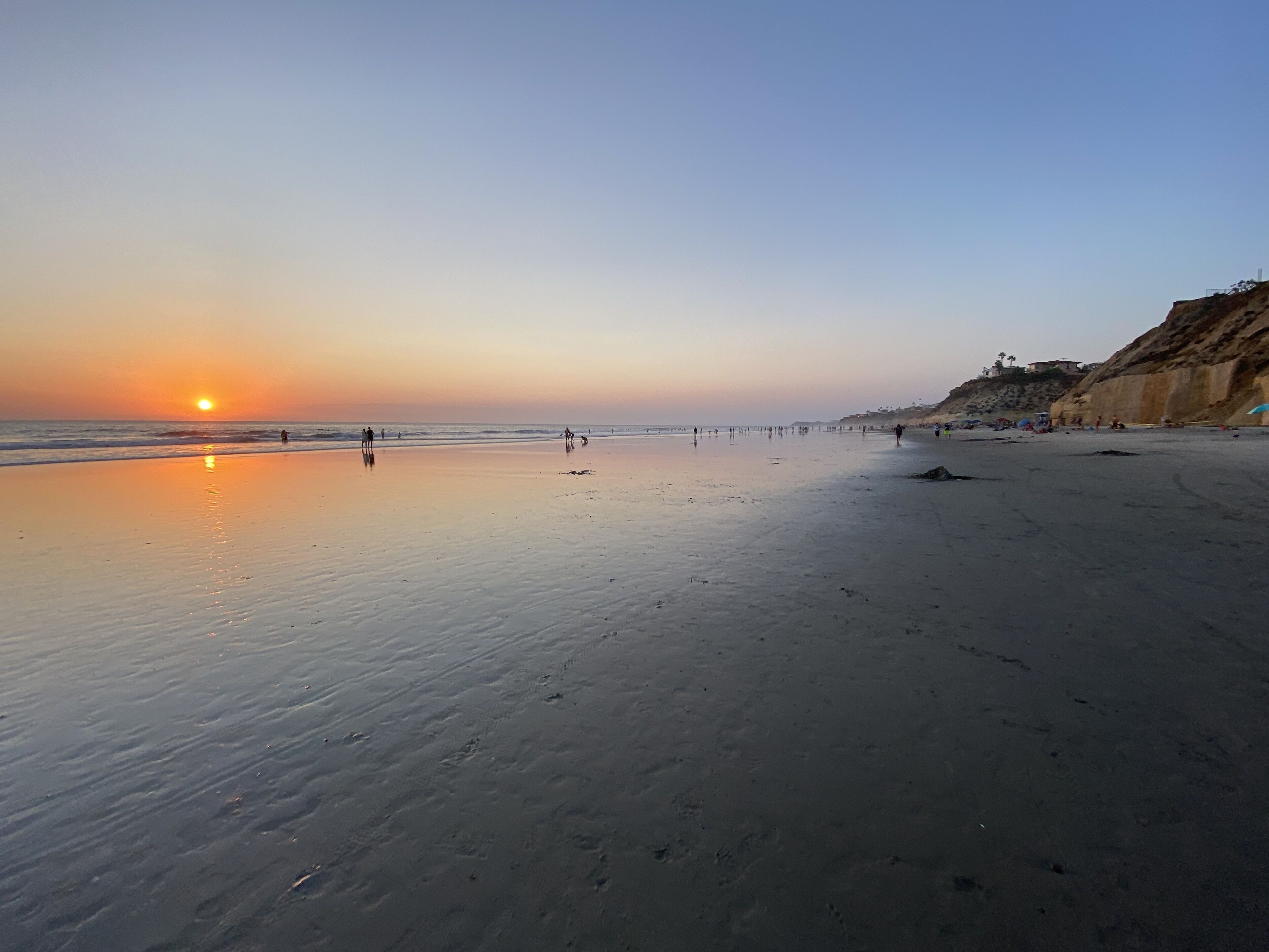 A beach with a sunset, calm sea, and a rocky cliff.