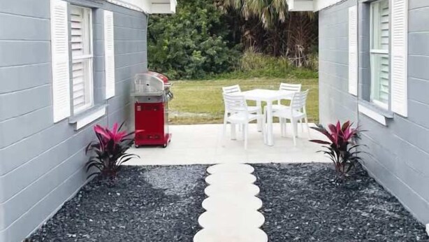Un espace patio avec une table et des chaises, un barbecue rouge et un chemin de jardin menant à une zone herbeuse.