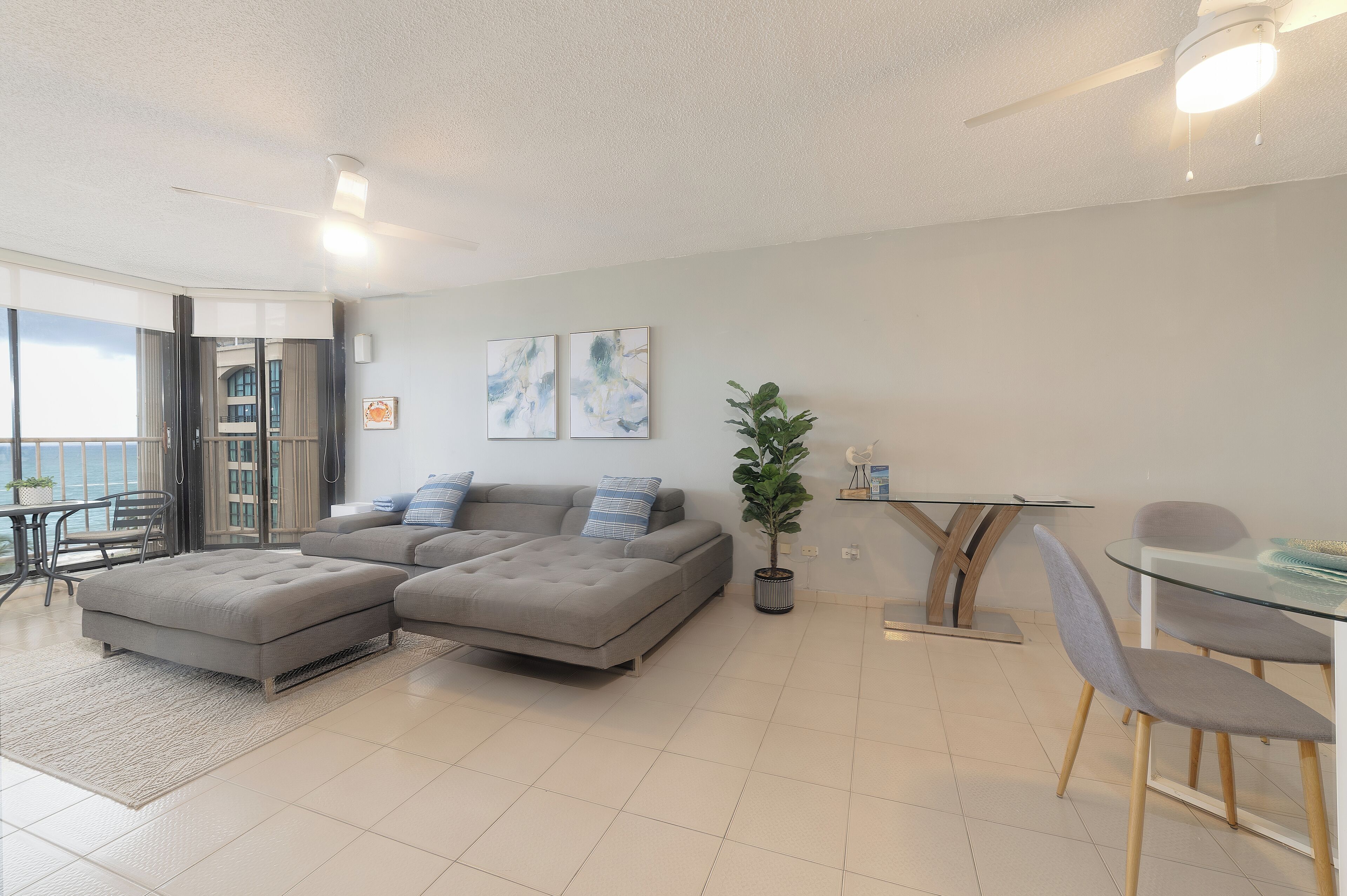 Een moderne woonkamer met een grijze bank, een poef en een glazen eettafel met stoelen. Er staat een potplant en er hangt kunst aan de muur.