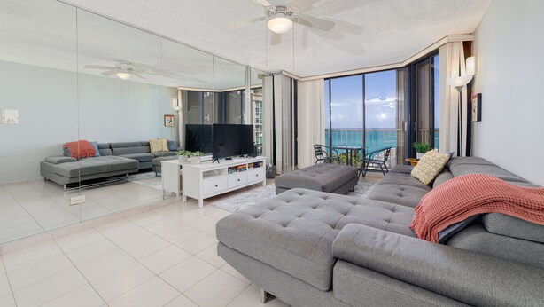 Un soggiorno moderno con un divano grigio, una TV a schermo piatto, una ventola a soffitto e un balcone con vista sul mare.