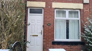 A red brick house with a white front door, a window with a black frame, and a small garden area with a metal gate. - Lovely 2-bed House in Bolton Greater Manchester (Bolton)