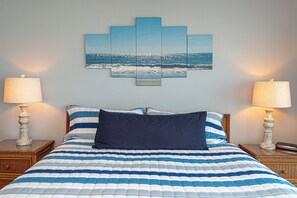 A bed with a striped blue and white comforter, two bedside lamps, a wall-mounted artwork of a beach scene, and a wooden headboard.