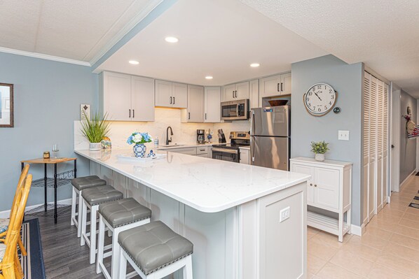 A modern kitchen with a central island, bar stools, stainless steel appliances, and a wall clock. - One of a Kind Direct Bay Views From This 3 Bedroom Condo at Assateague House! (Ocean City)