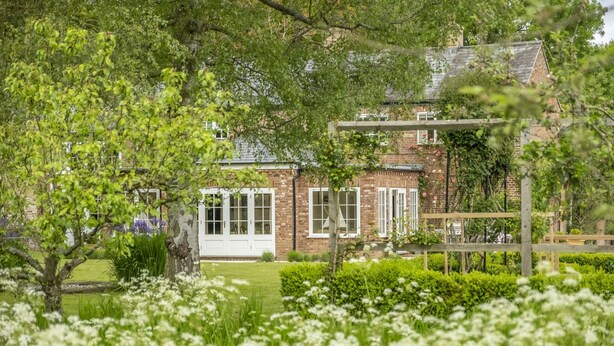 Una casa de ladrillo con contraventanas blancas, una pérgola de madera y un jardín con flores blancas.
