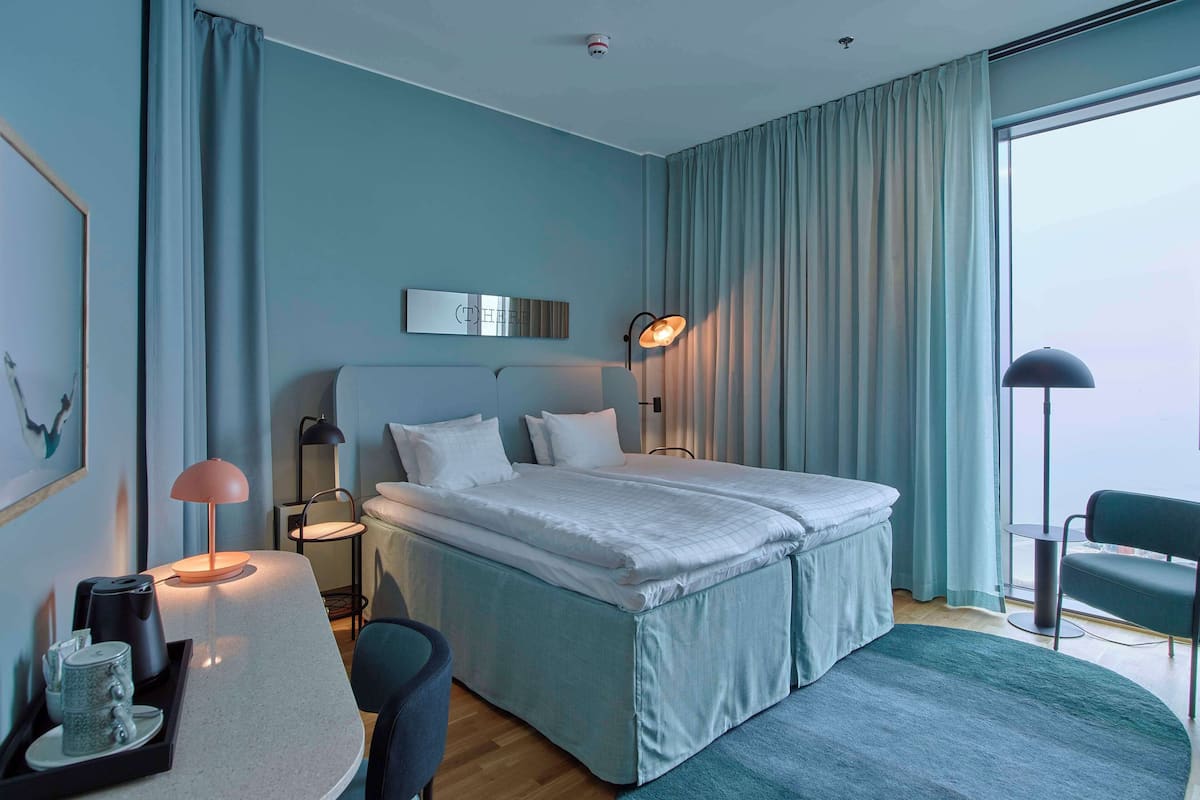 Habitación de hotel moderna con una cama grande, lámparas de noche, un escritorio con lámpara y vistas a la ciudad a través de amplios ventanales.