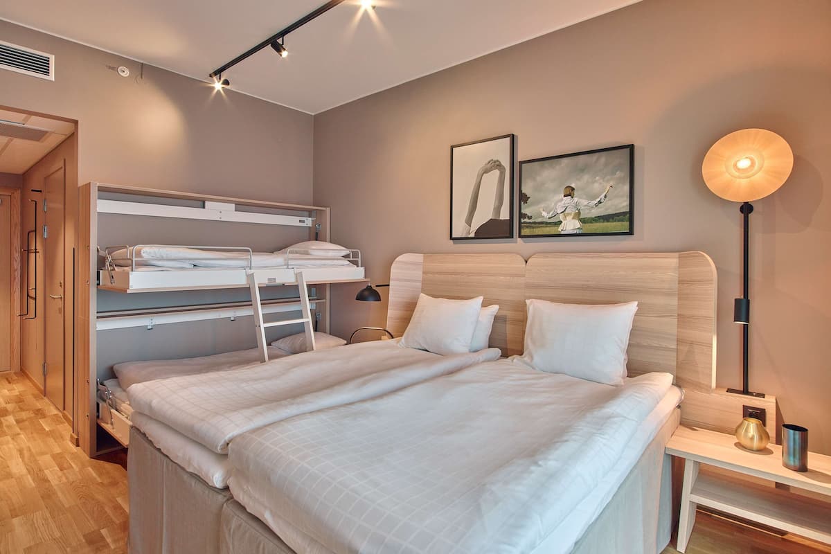 Habitación de hotel con literas, cama individual, mesita de noche con lámpara y dos cuadros en la pared.